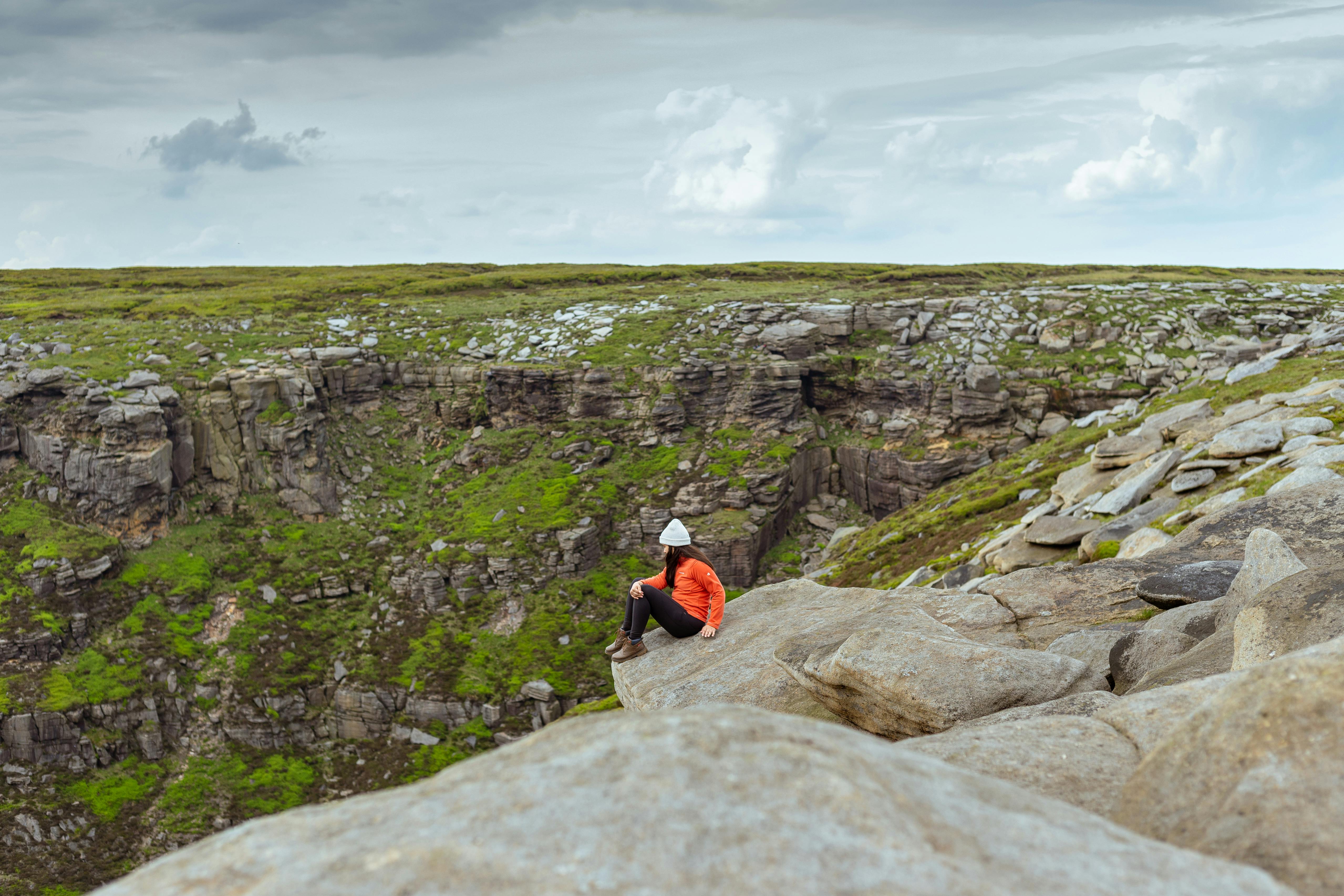 Person sitting on the edge of a rock formation at Stanage Edge, overlooking the rolling moors and valleys of the Peak District